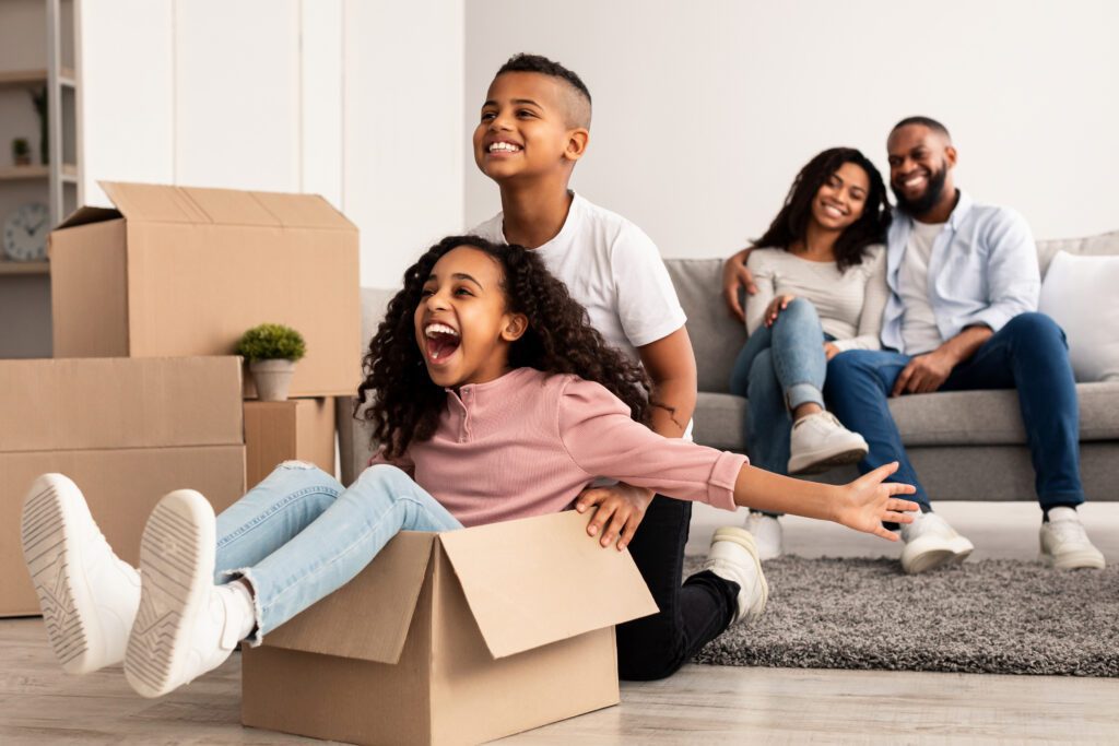 A girl in a cardboard box raises her hands to the sides while her brother pushes her like an airplane.