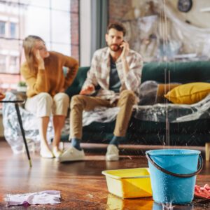 Water drips from the ceiling into a bucket with a couple in the background on the phone.