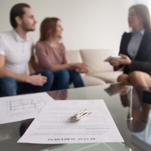 couple sitting on a couch with a realtor in the distance, a rental agreement in the forefront